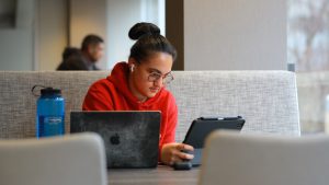 Student wearing red hooded sweatshirt looks at tablet with computer and water bottle also in front of her on table.