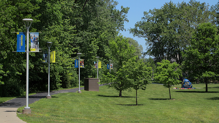 UToledo banners line the walkway along Ottawa River’s edge of the verdant Flatlands on Main Campus leading to Spirit Rock on a sunny spring day.
