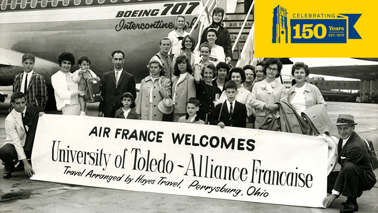 In this black-and-white photo for the late 1060s, members of The University of Toledo - Alliance Française pose for a photo on the tarmac after arriving in France for a cultural visit. They are holding a sign that says Air France Welcomes University of Toledo - Alliance Française Travel Arranged by Hayes Travel, Perrysburg, Ohio.