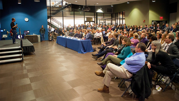 A photo of The University of Toledo Business Incubator's Pitch & Pour Competition with a presenter onstage addressing a small panel of judges and an audience of approximately 100 people.