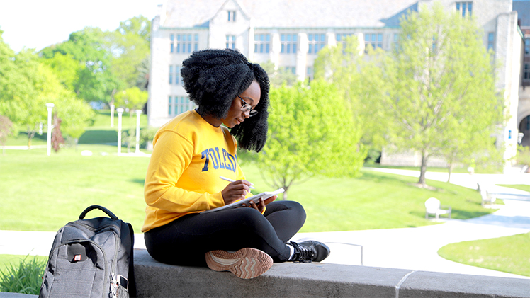 A UToledo student sits in the shade on a bench on Main Campus to read.