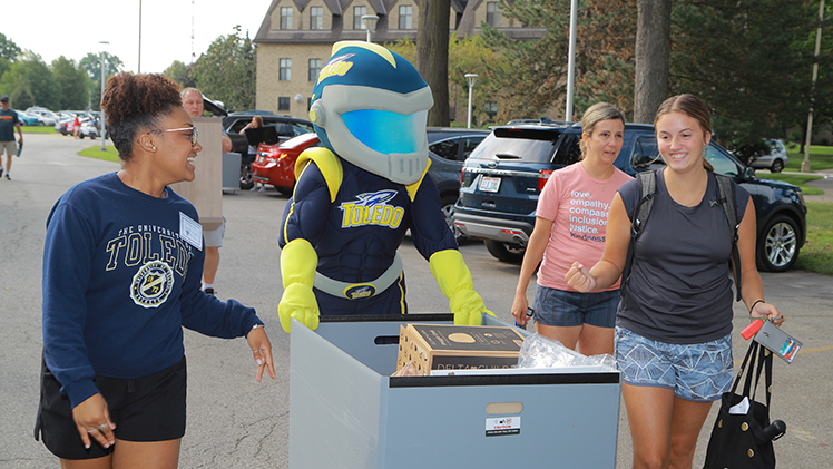 Rocky and a UToledo community campus volunteer help a new student move a cart full of boxes on a Move-In Day.