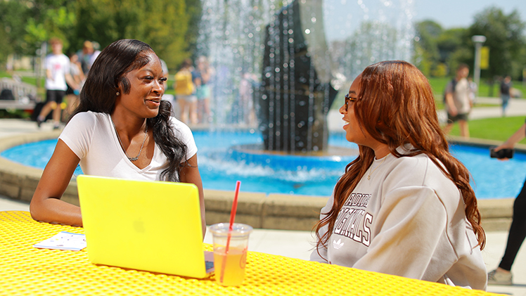 Two students talk in front of the fountain on UToledo Main Campus on a sunny day.