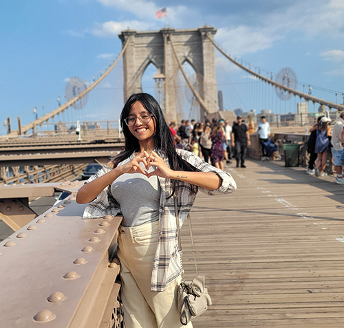 Feature photo of Swapnaa Balaji, a doctoral candidate in the College of Pharmacy and Pharmaceutical Sciences, making the heart sign with the Brooklyn Bridge in the background.
