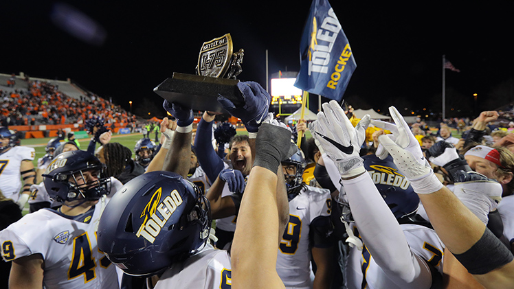 Rockets football team celebrates after beating BG, 32 to 31, Nov. 14, in a huddle holding the the Battle of I-75 trophy.