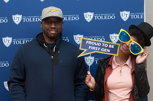 A man with a women, who is holding a "Proud to be First Gen" sign, pose for a photo during UToledo's annual celebration of first-generation students in the Thompson Student Union Ingman Room.