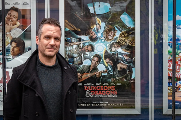 Photo of professional screenwriter, Michael Gilio, who cowrote the screenplay for the 2023 film, “Dungeons & Dragons: Honor Among Thieves,” standing in front of the film's movie poster at a movie theater.