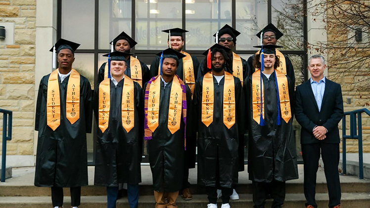 UToledo Football Head Coach Jason Candle poses with graduating players in commencement cap and gowns.