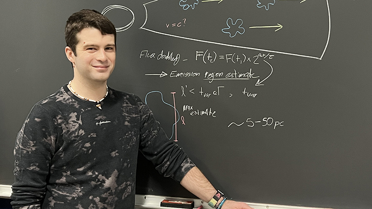 Photo of Tyler Robbins, a senior pursuing an astrophysics degree with a minor in mathematics, posing in front of a black board wiht an equation written on it.
