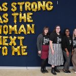 Four women pose in front of a words on a wall: As strong as the woman next to me.