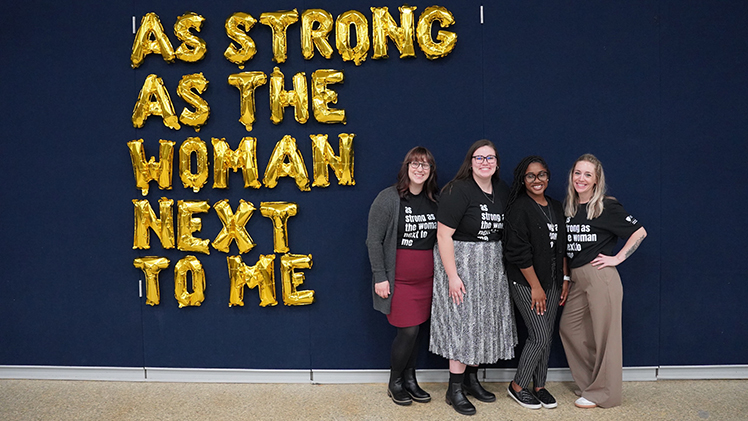 Four women pose in front of a words on a wall: As strong as the woman next to me.
