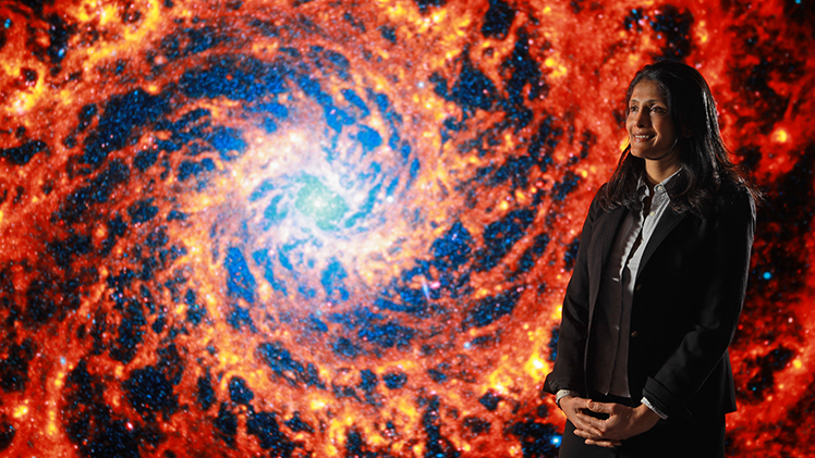 Photo of Dr. Rupali Chandar, a professor and associate chair of the Department of Physics and Astronomy, standing in front a giant image of fire-red galaxy. 