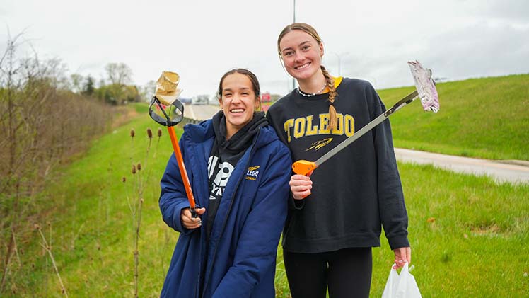 2 female UToledo student-athletes pose as they clean up the Toledo community on 419 Day.