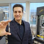 Dr. Meysam Haghshenas, an assistant professor in the Department of Mechanical, Industrial and Manufacturing Engineering, holds an hourglass test specimen in front of the new high-temperature ultrasonic fatigue tester in the the Failure, Fracture and Fatigue Laboratory.