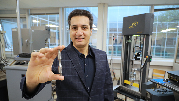 Dr. Meysam Haghshenas, an assistant professor in the Department of Mechanical, Industrial and Manufacturing Engineering, holds an hourglass test specimen in front of the new high-temperature ultrasonic fatigue tester in the the Failure, Fracture and Fatigue Laboratory.