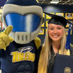 Business Gradate Student Molly Bennett, dressed in graduating cap and gown and holding a diploma, poses with UToledo mascot Rocky. 