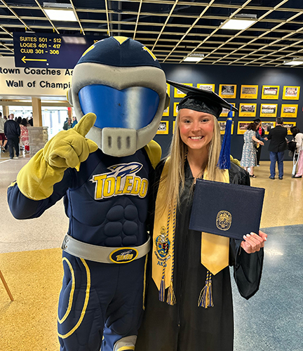 Business Gradate Student Molly Bennett, dressed in graduating cap and gown and holding a diploma, poses with UToledo mascot Rocky. 