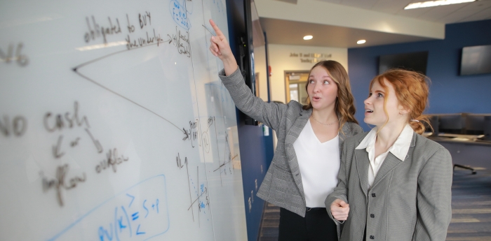 Two women point to math equations written on a white board.