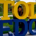 Photo of graduating UToledo student posing with her head in the "O" of the Tol-Edo sign.