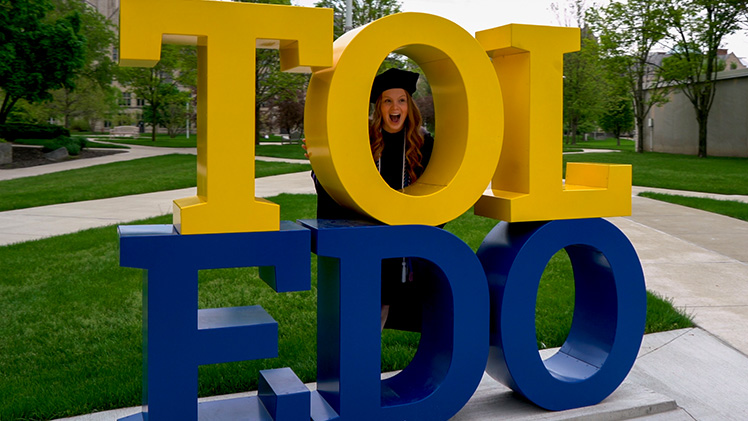 Photo of graduating UToledo student posing with her head in the "O" of the Tol-Edo sign.