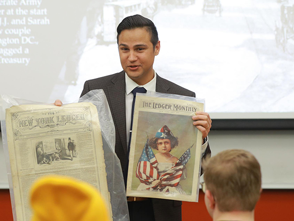 Photo of Dr. Ayendy Bonifacio, an assistant professor of English specializing in U.S. ethnic literary studies, holding old newspapers in a classroom as he teaches students.