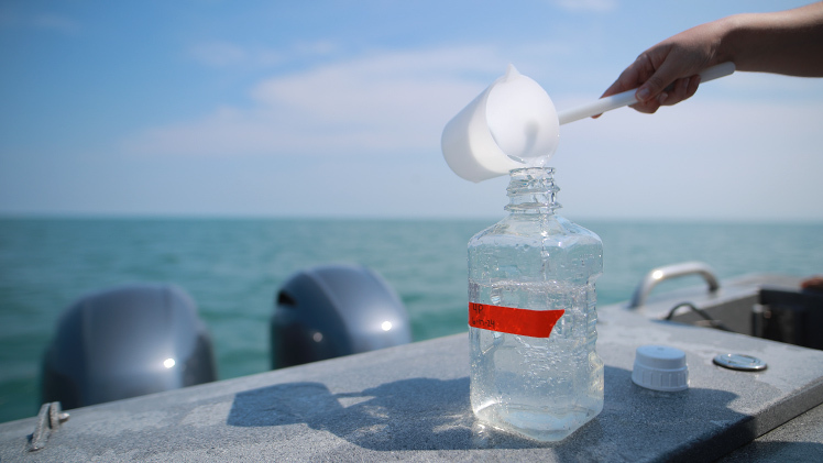 A photo taken while on Lake Erie of a hand as it pours water from a cup into a large glass jar.