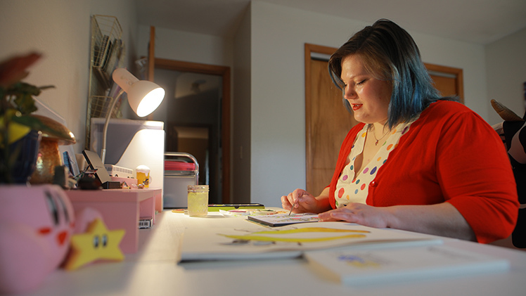 Zoey Maness, 28, a junior studying communication who works full time at UTMC as a staffing coordinator for nursing administration, works on a watercolor painting at her desk.