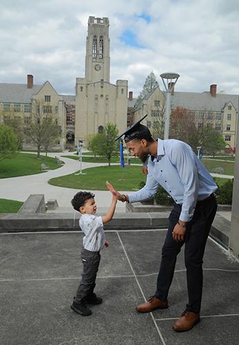 A UToledo student parent wearing a graduation cap high-fives his young son on Centennial Mall.