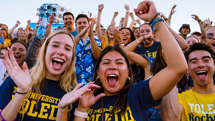 A group of UToledo students smile and cheer during a Rockets football game.