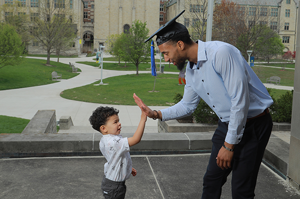 A UToledo student parent wearing a graduation cap high-fives his young son on Centennial Mall.