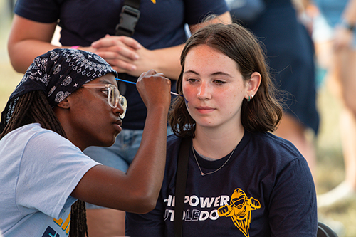 A female UToledo student paints the face of another female student at the annual CAP Carnival at Carter Field as part of the 2024 Rocket Welcome Weekend.