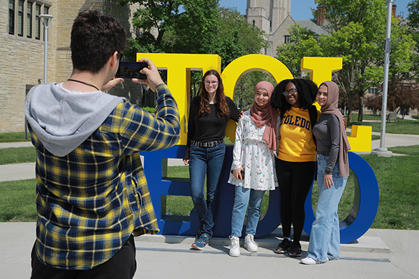 A student takes a photo of four students posing by the TOL-EDO sign