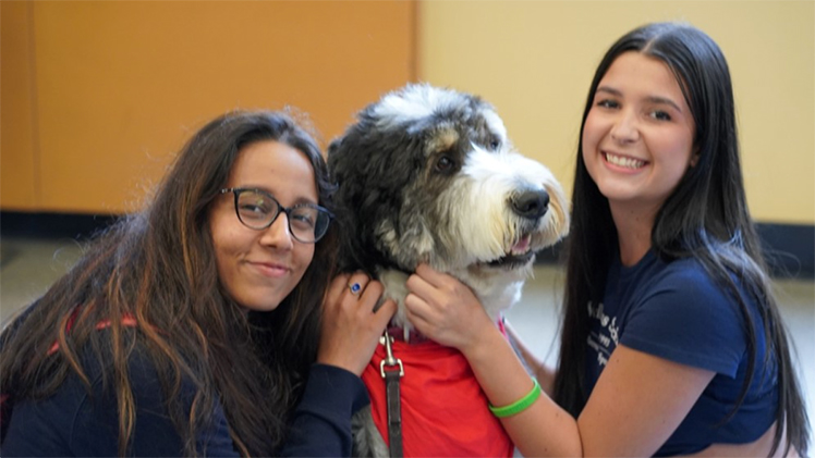 Two female UToledo students smile for the camera as they pet a dog at a Paws on Campus event.