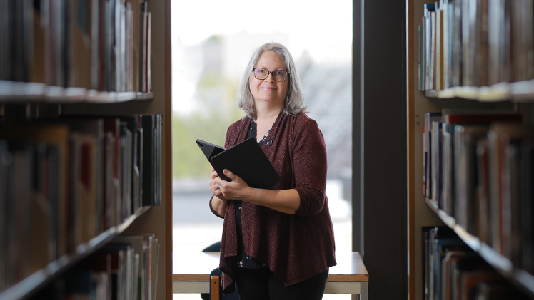 Portrait of Lucy Duhon is an associate professor and scholarly communications librarian for University Libraries, who is holding a book in a library.