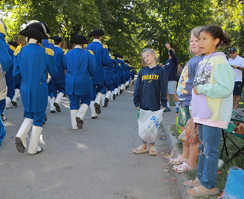 Three young girls watch members of the parade dressed in costumes from Colonial-era America march by.