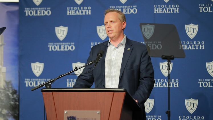 UToledo Interim President Matt Schroeder speaks from behind the podium in the Student Union in front of a blue backdrop with white UToledo and UToledo Health logos repeating across it.