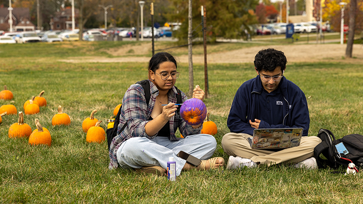 Tejal Thakur, a junior studying legal and paralegal studies, paints a pumpkin as Amaan Boghani, a junior studying communication, studies on his laptop.