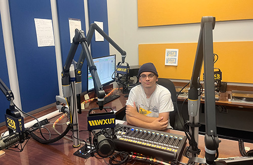 Photo of Trip Floyd, a junior studying communication, sitting in the WXUT radio station studio surrounded by the station's equipment.