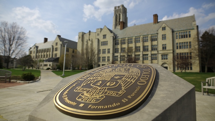 Photo with University Seal in Centennial Mall in the foreground with University Hall behind it under a blue sky.