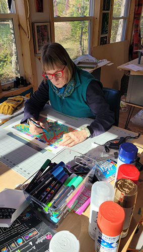 Photo of Barbara Miner, professor and chair of the Department of Art, working on her colorful new signal box wrap design at a desk in her home studio.