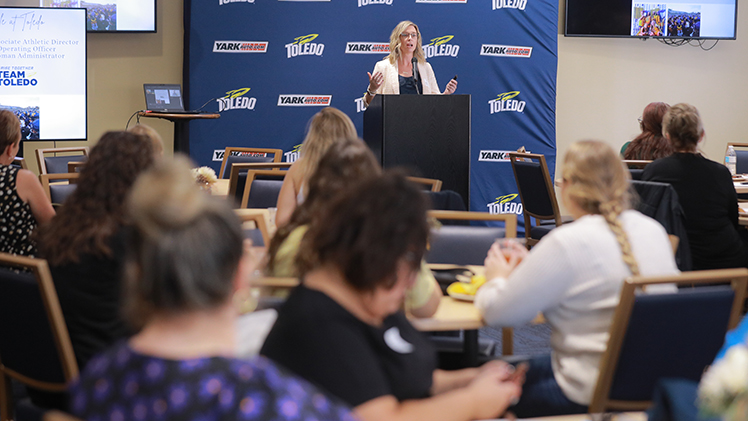 Photo of Nicole Alderson at a podium during a University of Women's Commission luncheon event.