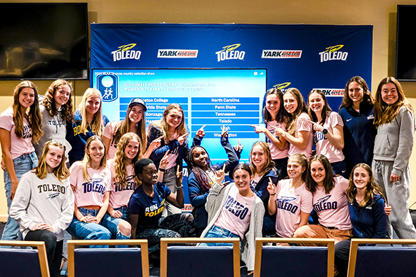 Group photo of the Toledo women’s cross country celebrating in front of a giant screen announcing that the team earned an at-large berth to Saturday's national title race.