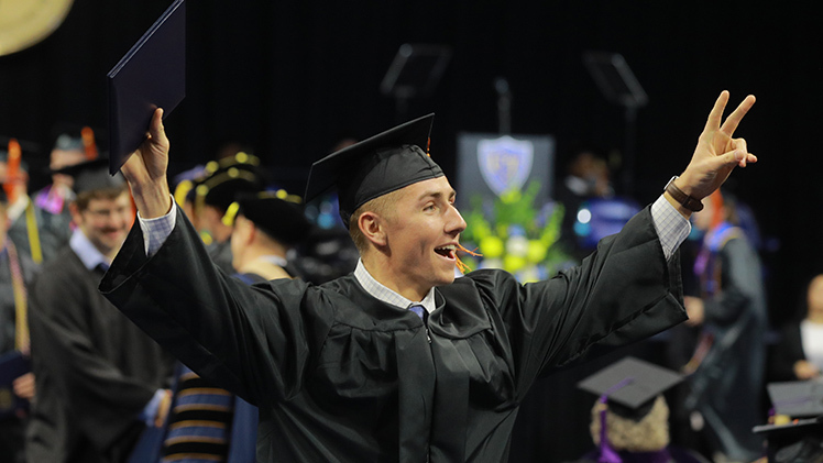 A male student holds his diploma high in one hand and makes the peace sign with the other hand during UToledo's 2024 fall commencement ceremony.