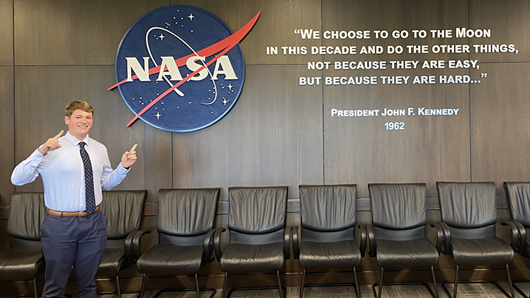 Photo of Reed Schumacher standing in an office at NASA Kennedy Space Center pointing to a NASA sign on the wall with a quote from Pres. John F. Kennedy: “We choose to go to the moon in this decade and do the other things, not because they are easy, but because they are hard … “