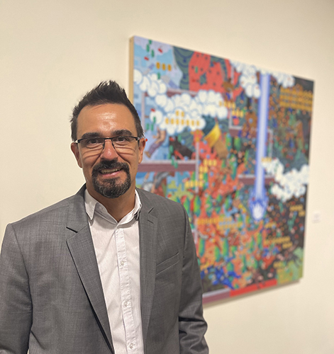 Portrait of Dan Hernandez next to one his paintings. He teaches drawing and painting as a professor in the Department of Art.