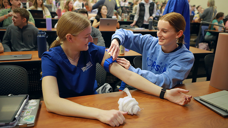 Abi Ovacek (Senior majoring in Nursing) watches as Elyse Bell (Senior majoring in Nursing) practices the proper way to apply a tourniquet during the Stop the Bleed event.