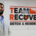 Matt Bell, who will graduate May 2 with his degree in social work, stands in front of a Team Recovery sign.