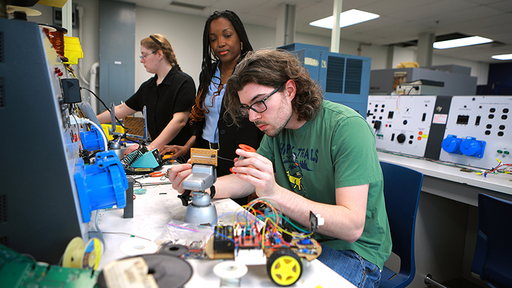 Dr. Ngalula Sandrine Mubenga, center, an associate professor in the Department of Engineering Technology, looks on as Ethan Ritchey, right, an electrical engineering technology junior solders a transistor, in the foreground. Sarah Cook, an electrical engineering technology senior, is the foreground.