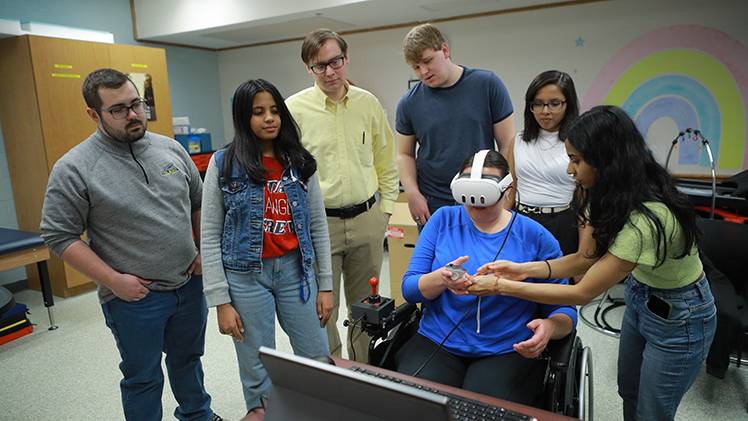 A team of undergraduate seniors developed a virtual reality tool to assess whether a person can safely maneuver a motorized wheelchair in collaboration with Sunshine Communities. From left, Evan Palmer, Simran Shekhar, Jeffrey Walter of Sunshine Communities, Nick Hemmert, Tika KC and Sakshi Dhumma watch as Sunshine’s Kelsey Garner tries it out at the nonprofit in Maumee.