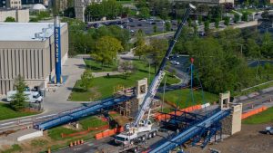 A drone shot shows the construction site for UToledo's new footbridge over Douglas Road from above, as a crane moves one of the bridge sections into place.